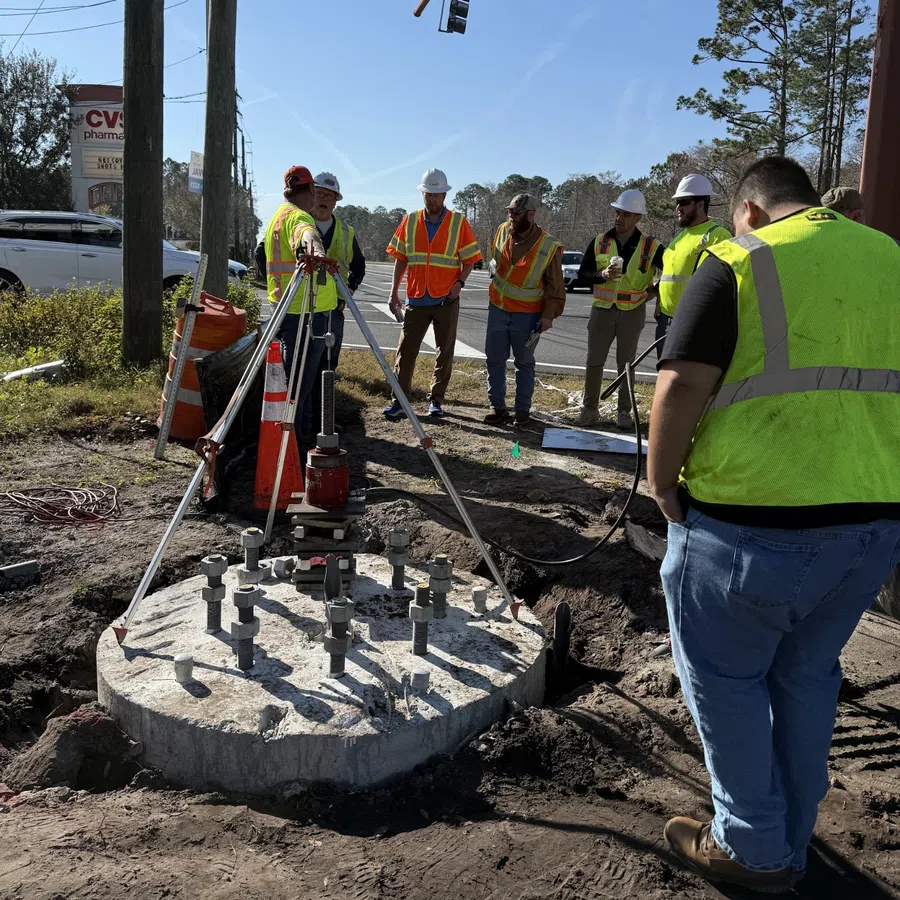 Construction workers in safety vests and helmets inspect concrete foundation with metal bolts by roadside under traffic lights.