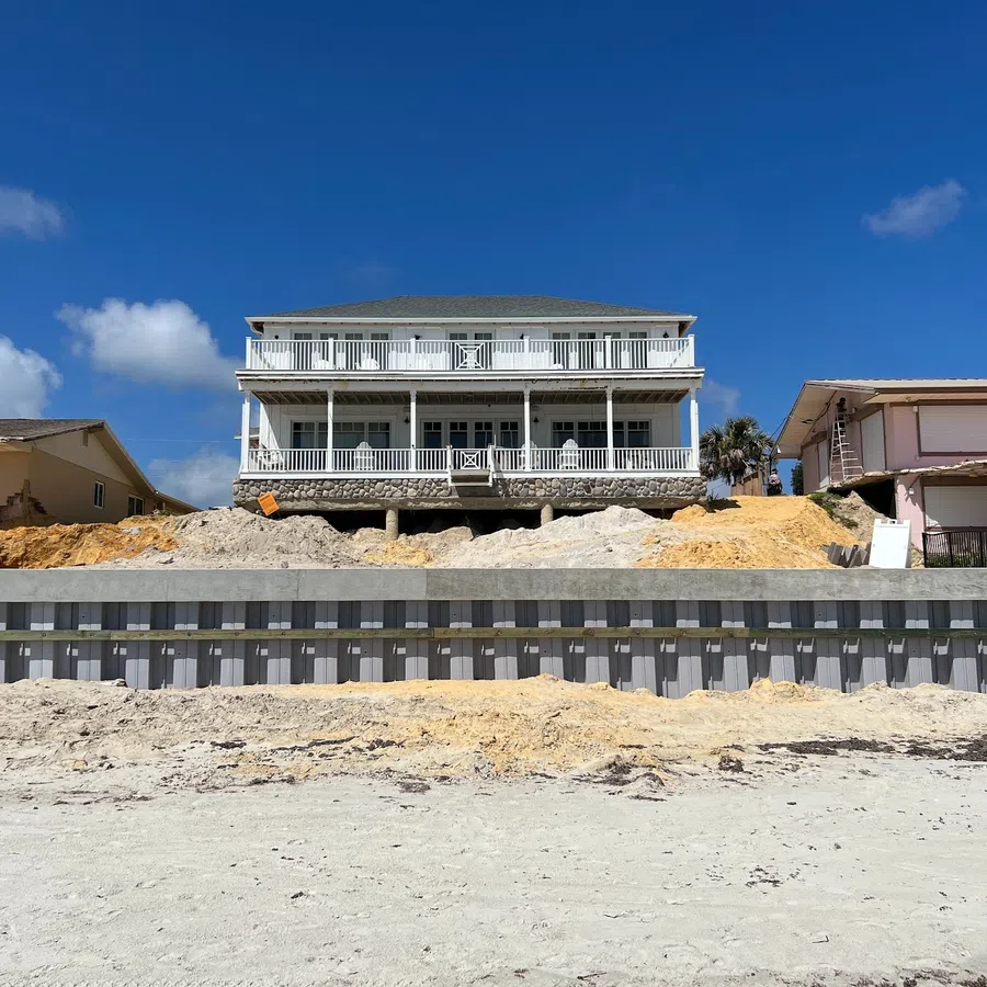 Two-story white beach house elevated on stilts with sandy shore and seawall under clear blue sky.