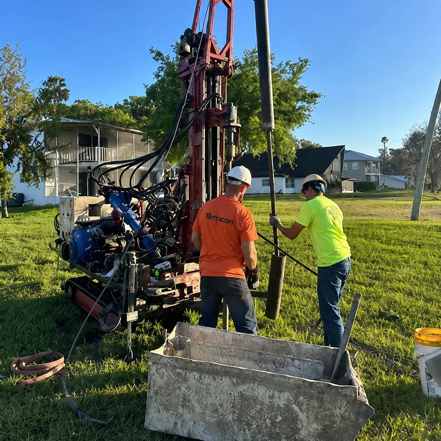 Two workers operating a drilling rig outdoors with one lifting equipment in a sunny residential area.