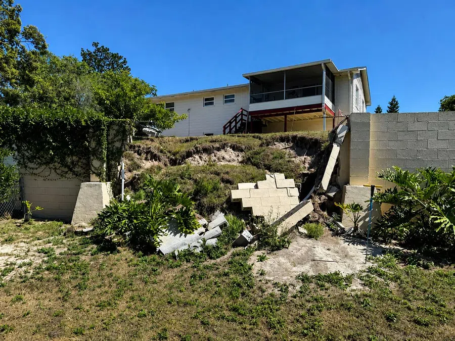 Damaged retaining wall collapsed near grassy backyard slope beneath a house with blue sky above.