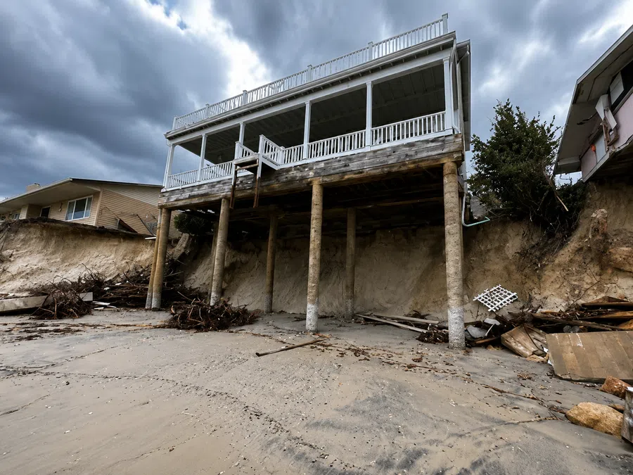 Beach house elevated on stilts with visible erosion under the structure and debris on the sandy shore.