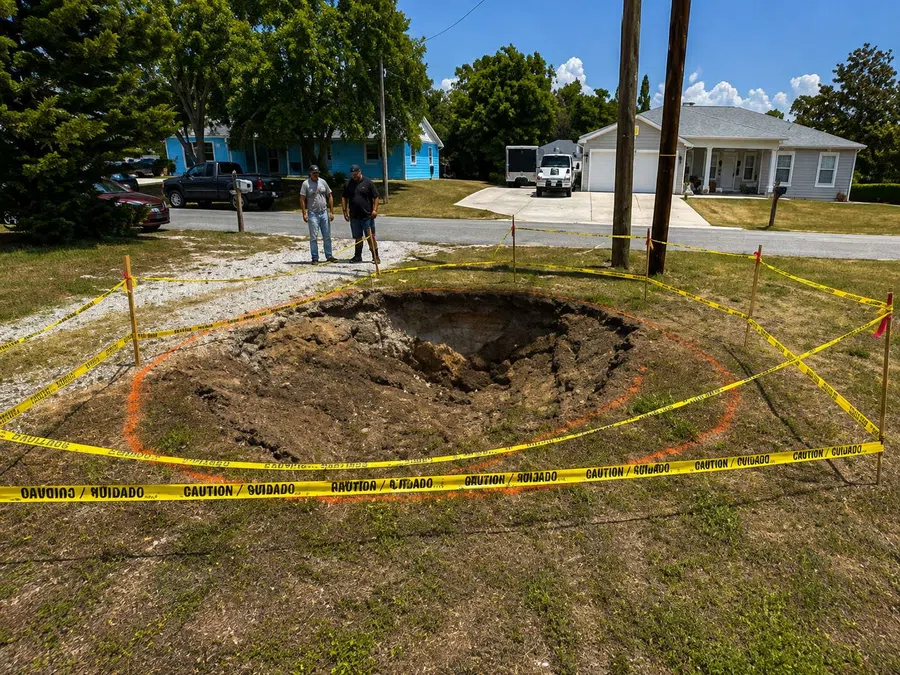 Large sinkhole in a residential yard surrounded by yellow caution tape with two men observing nearby.