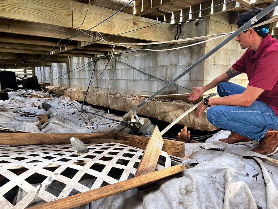 Man inspecting damaged wooden beams and debris under a raised house with exposed foundation.