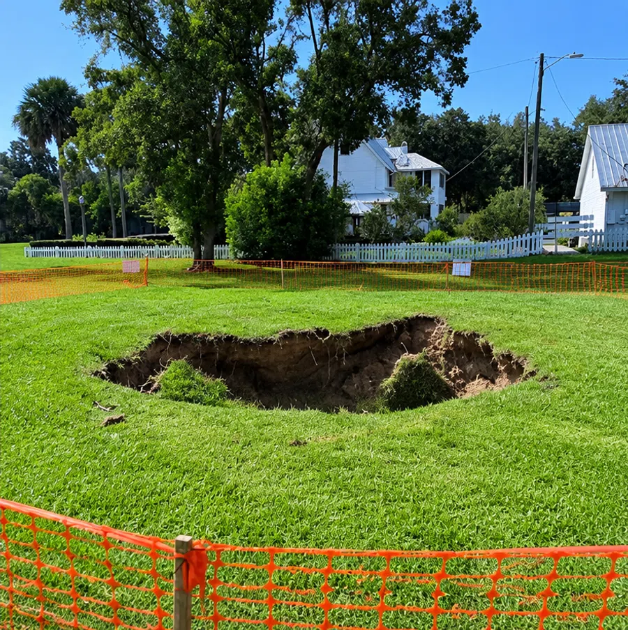 Large sinkhole in grassy area surrounded by orange safety fencing near white houses and trees.