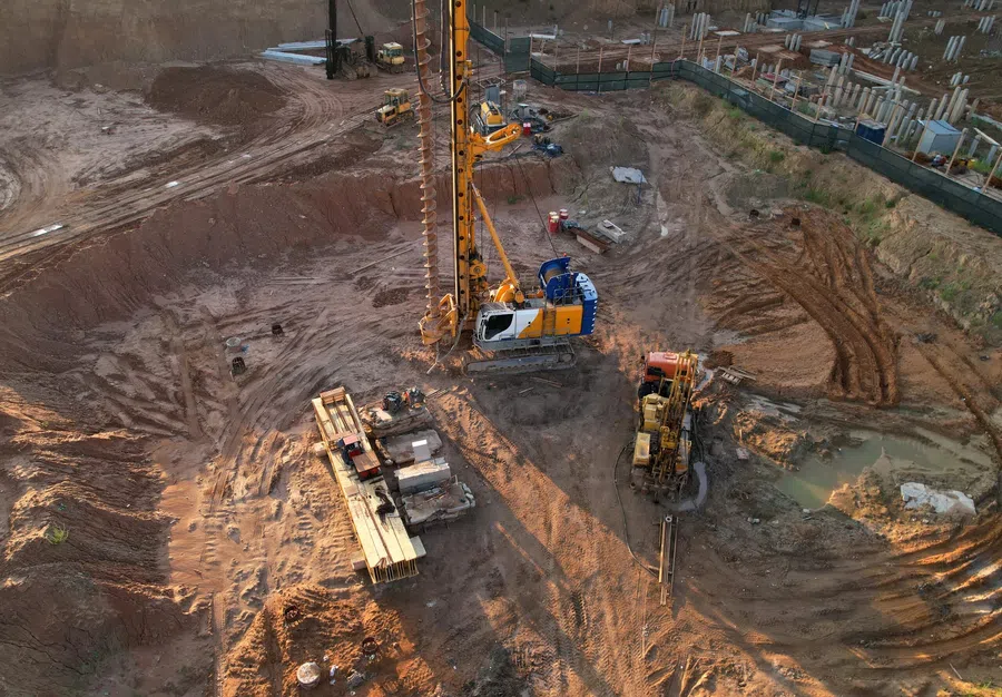 Aerial view of a construction site with heavy machinery drilling into the ground and building foundations nearby.