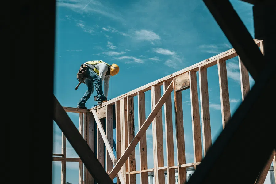 Construction worker in safety gear working on wooden framework of a building under clear blue sky.