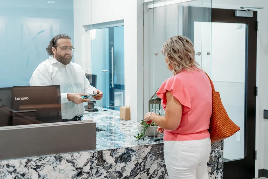 Woman at a reception desk talking to a man behind a protective glass in a modern office setting