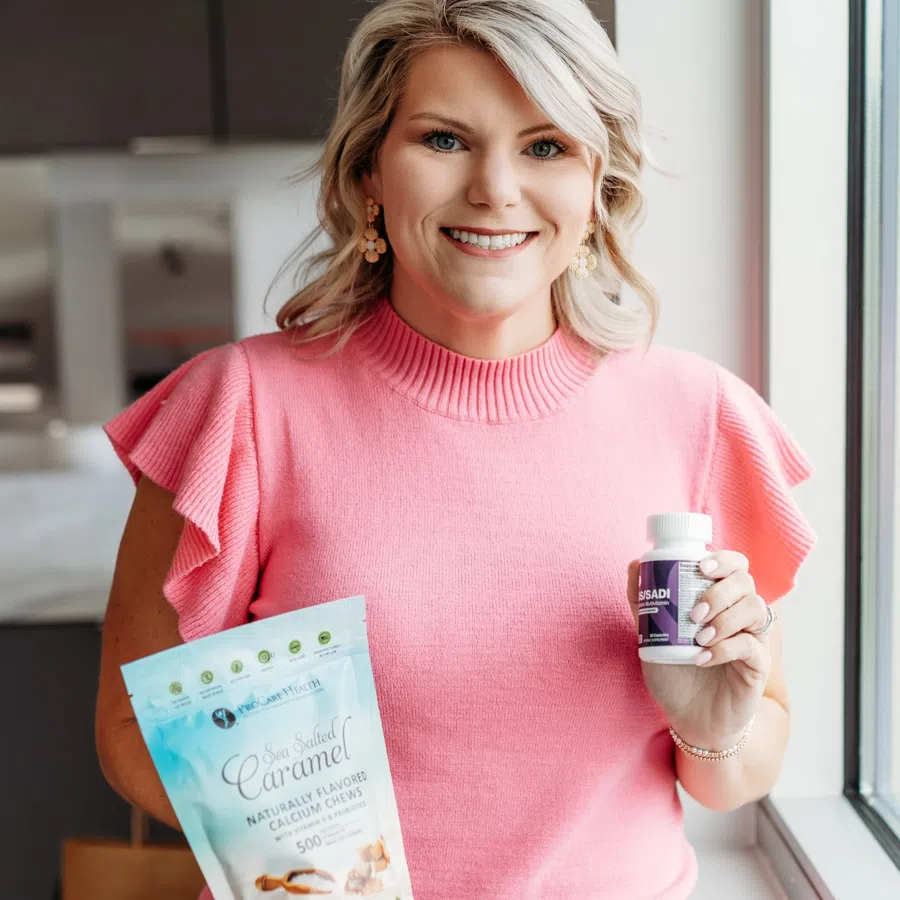 Woman in pink sweater holding sea salted caramel calcium chews and a supplement bottle by a window.
