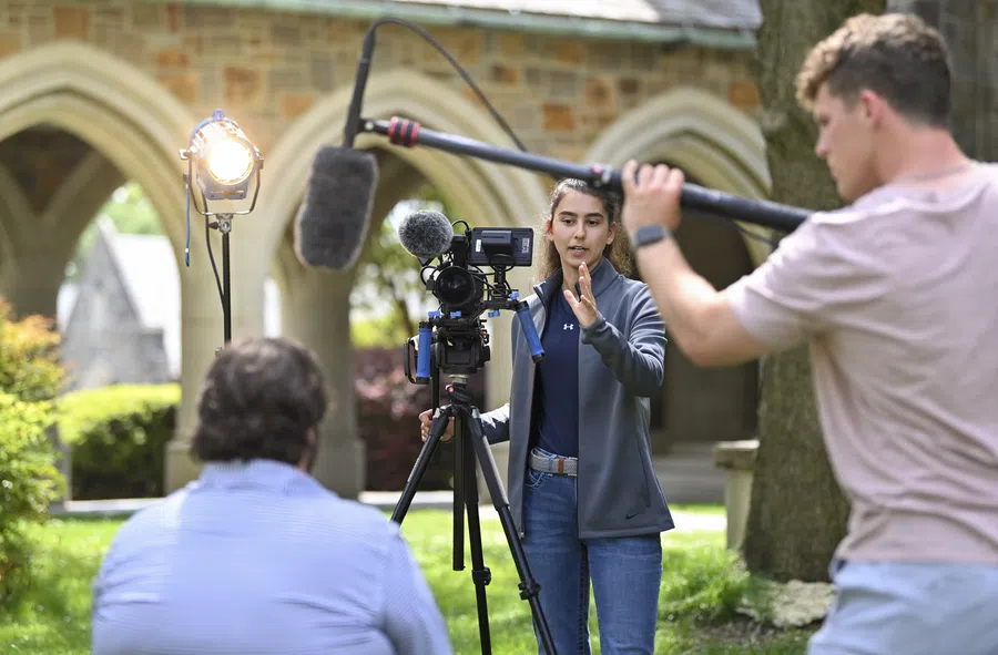 Film crew recording outdoors with camera, boom mic, and lighting in a garden with stone arches in background