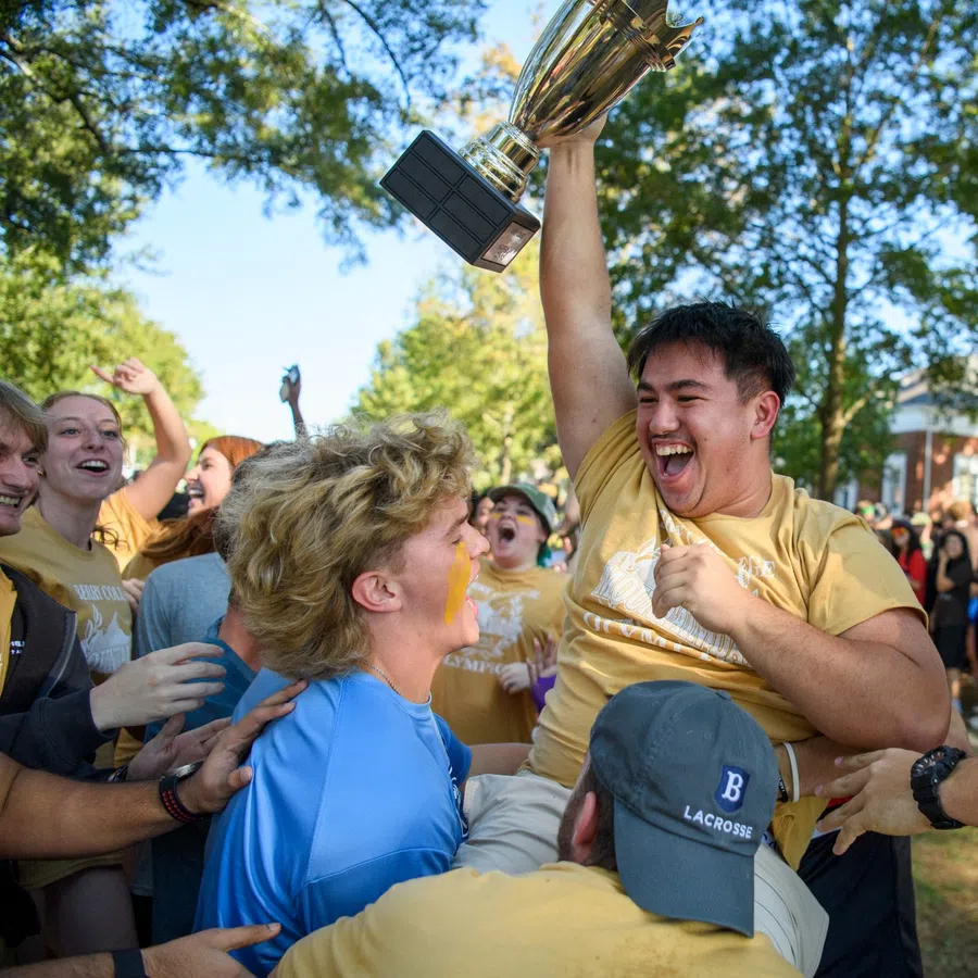 Group of college students celebrating as one holds a trophy high outdoors with trees and sunlight.