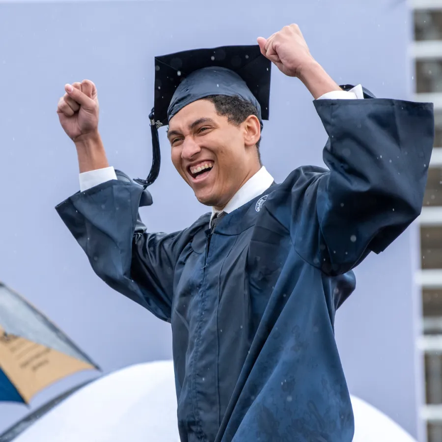 Happy male graduate wearing cap and gown celebrating achievement outdoors in the rain with raised fists