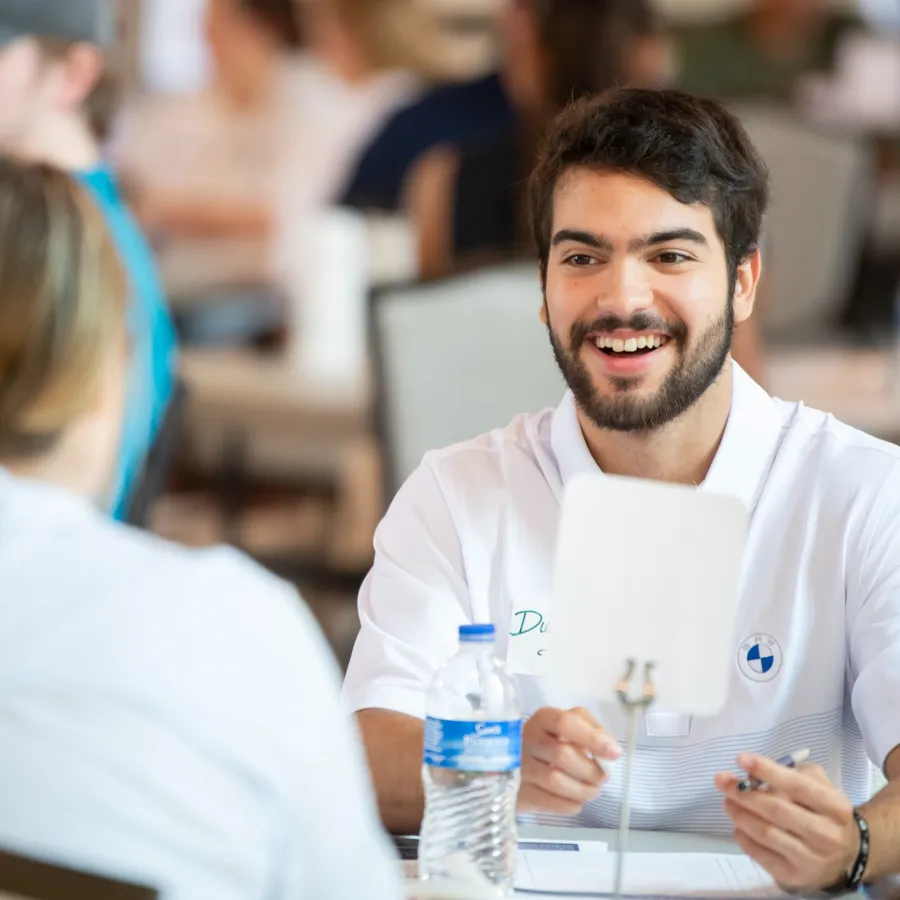 Smiling young man in white shirt engaging in conversation at a busy indoor event with a water bottle on the table.