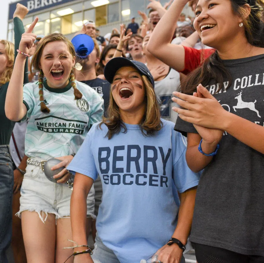 Young fans cheering energetically at a Berry College soccer game with face paint and team shirts.