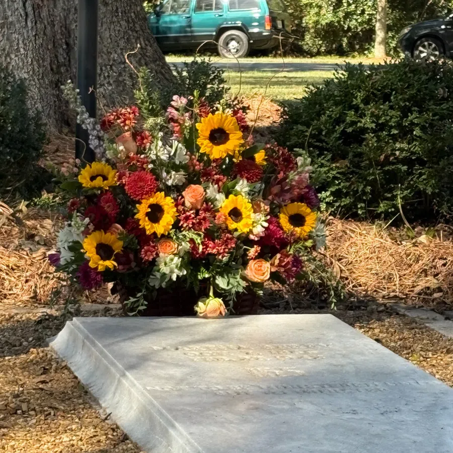 Colorful bouquet of sunflowers, roses, and other flowers placed at a grave in a peaceful outdoor setting.