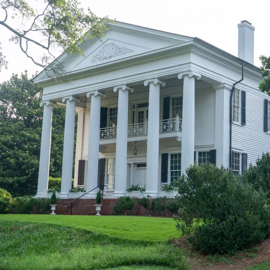 Large white plantation-style house with tall columns, black shutters, and lush green lawn under clear sky.