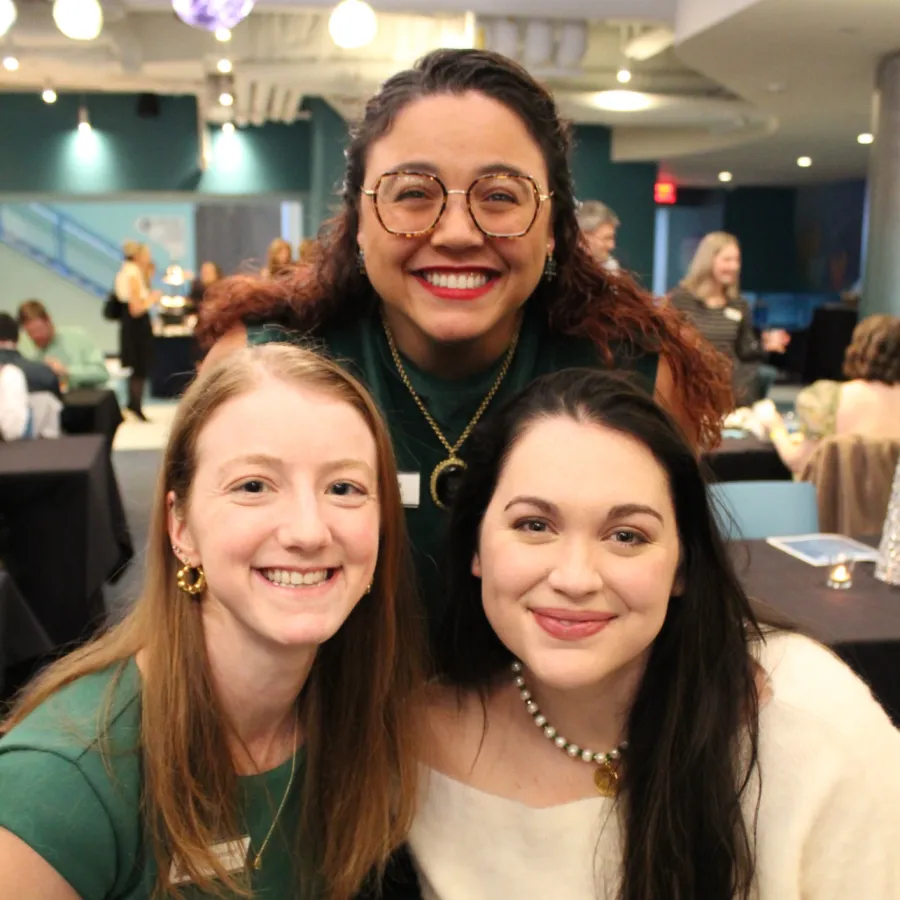 Three women smiling at a social event indoors with tables and people in the background.