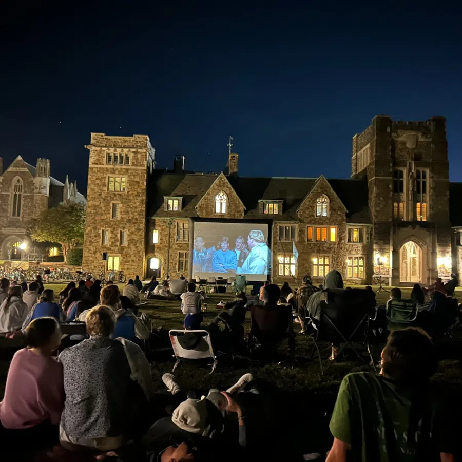 Crowd watching outdoor movie screening at night on lawn in front of historic stone building with lit windows.