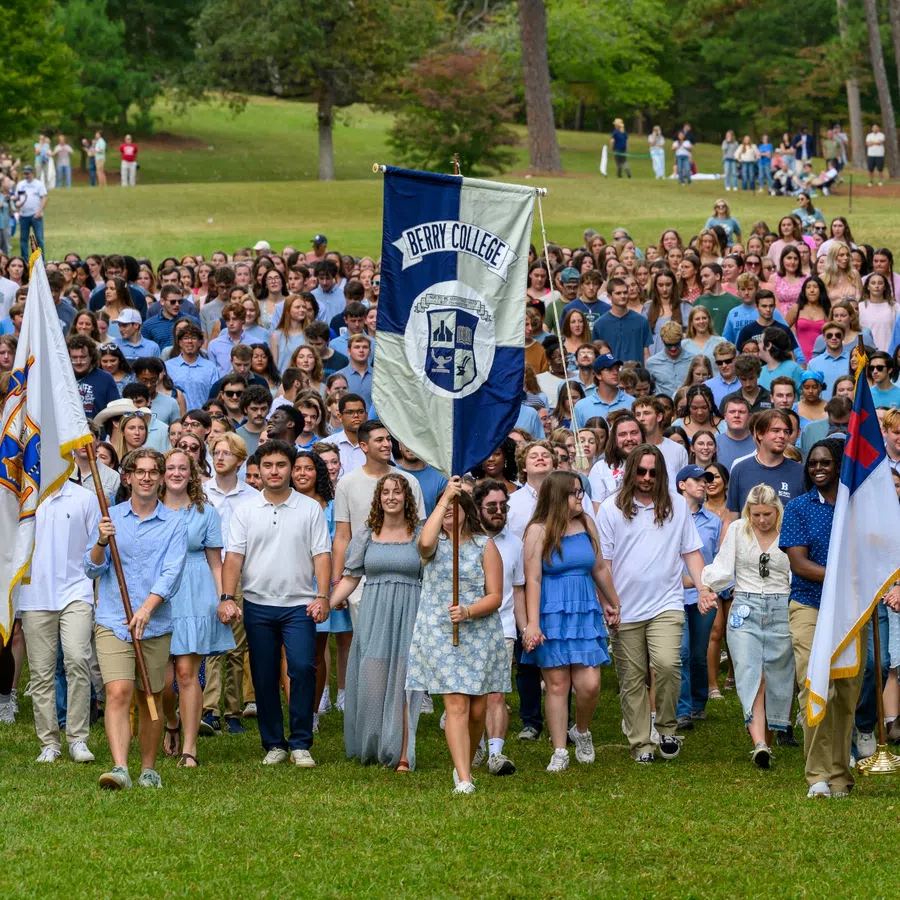 Large group of Berry College students walking outdoors on grass holding college banners during event.