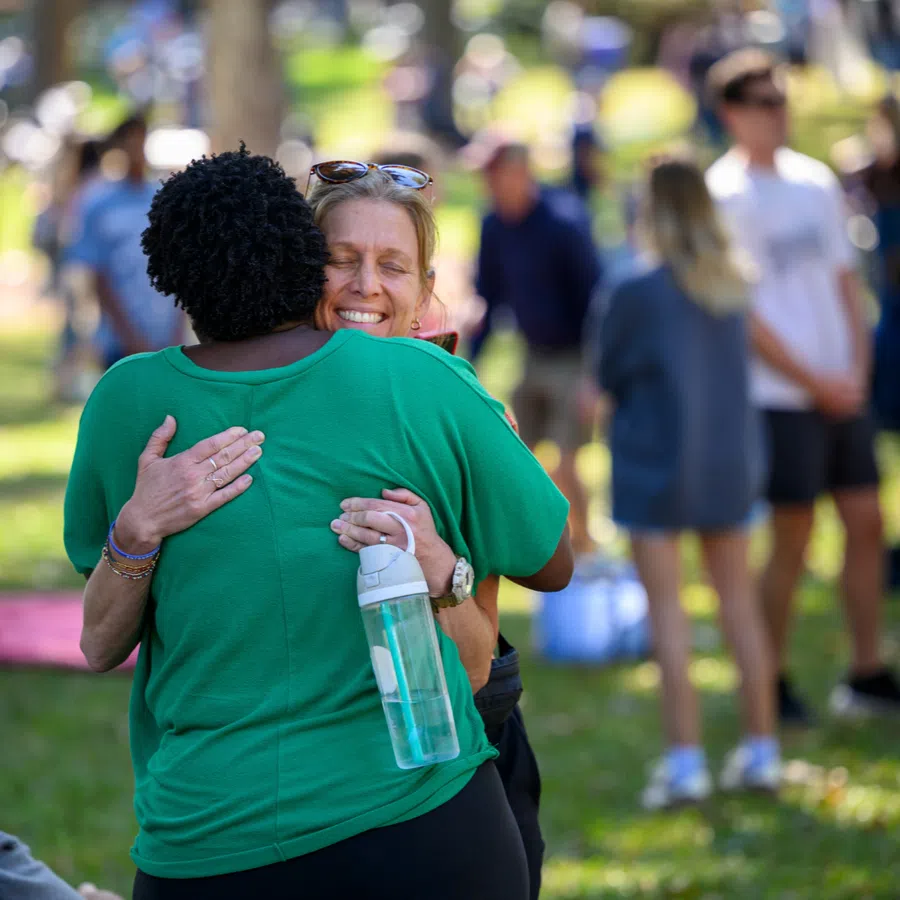 Two women warmly hugging outdoors at a sunny park with people mingling in the background.