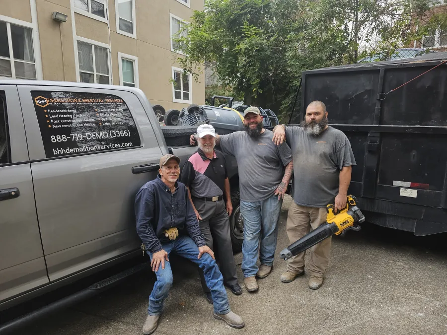 Four demolition workers posing by a truck and dumpster, one holding a yellow leaf blower outdoors in an urban area.