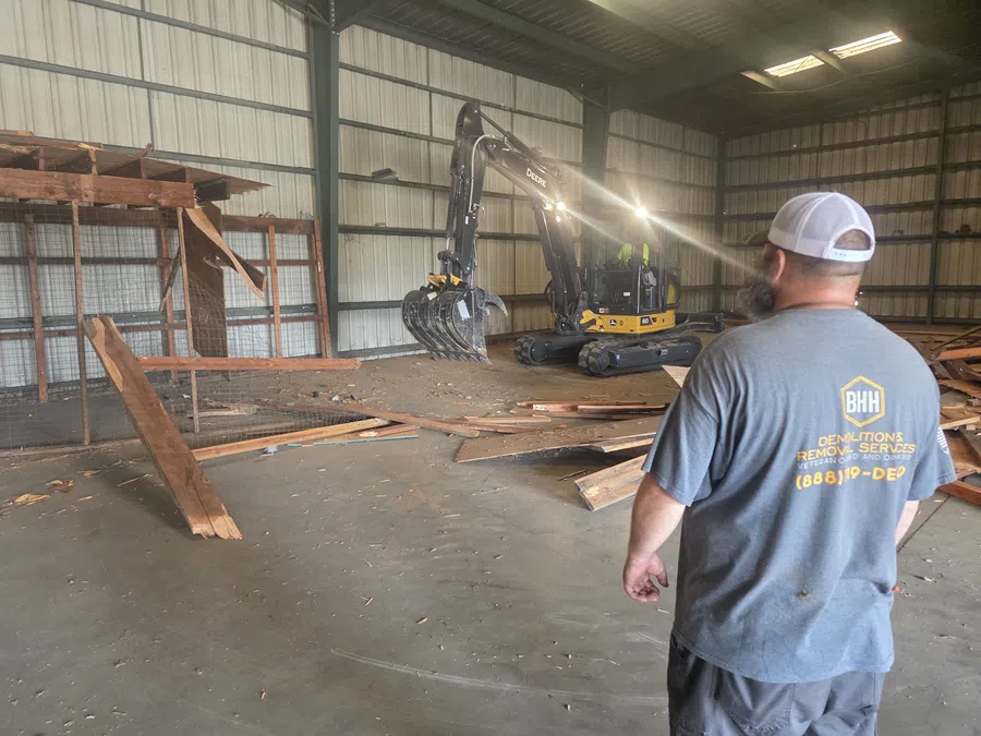 Worker in gray shirt overseeing indoor demolition with excavator clearing wooden debris inside metal building.