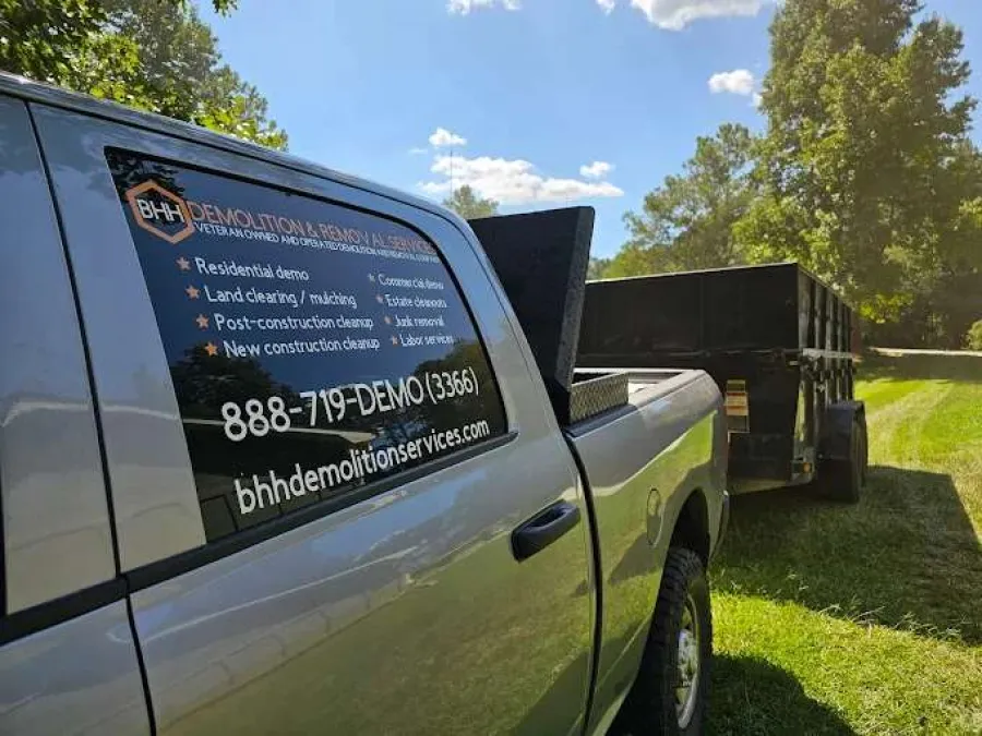 Gray pickup truck with BH Demolition & Removal Services advertisement towing a trailer on green grass under a blue sky.