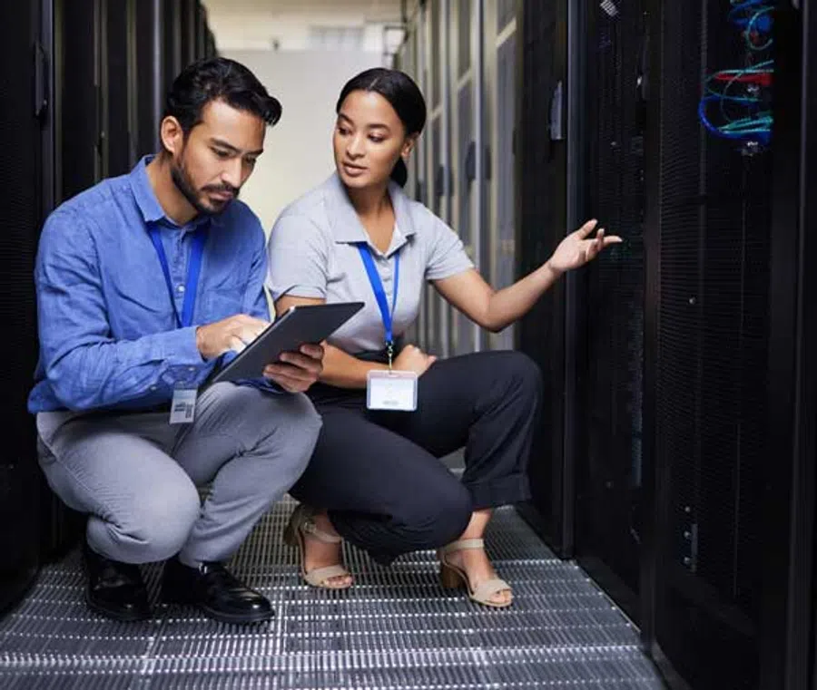 Two IT professionals wearing ID badges work together in a data center server room with digital tablet and racks.