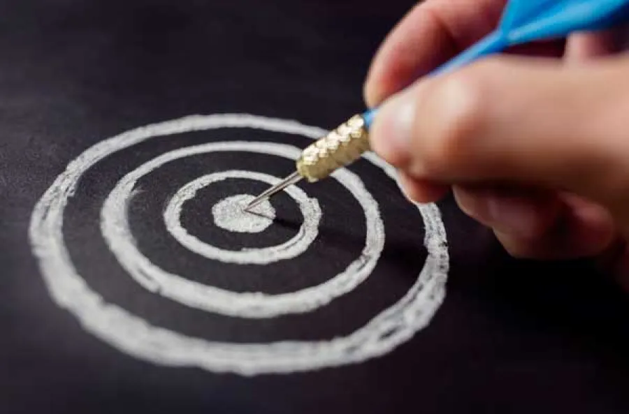 Hand holding a blue dart aiming at the center of a white chalk-drawn target on a blackboard surface