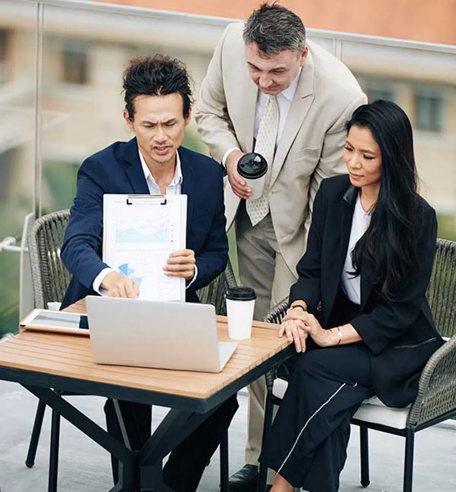 Three business professionals reviewing financial charts on a laptop and clipboard during an outdoor meeting.