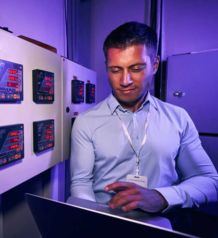 Engineer in blue shirt working on laptop near control panel with illuminated digital meters in dimly lit room.