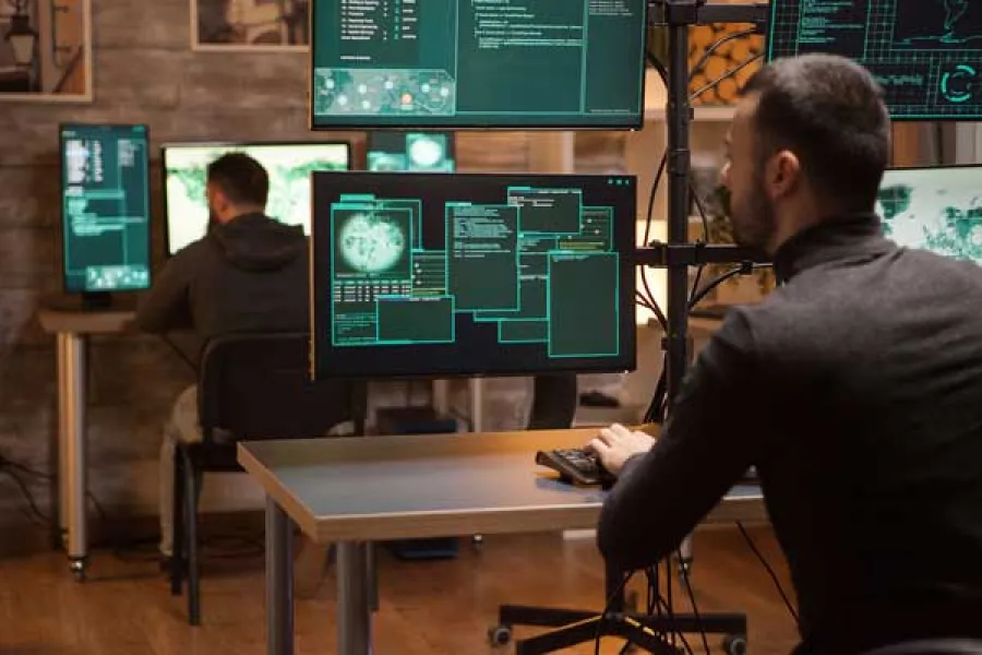 Two men working on multiple computer screens displaying data and cybersecurity code in a tech office.