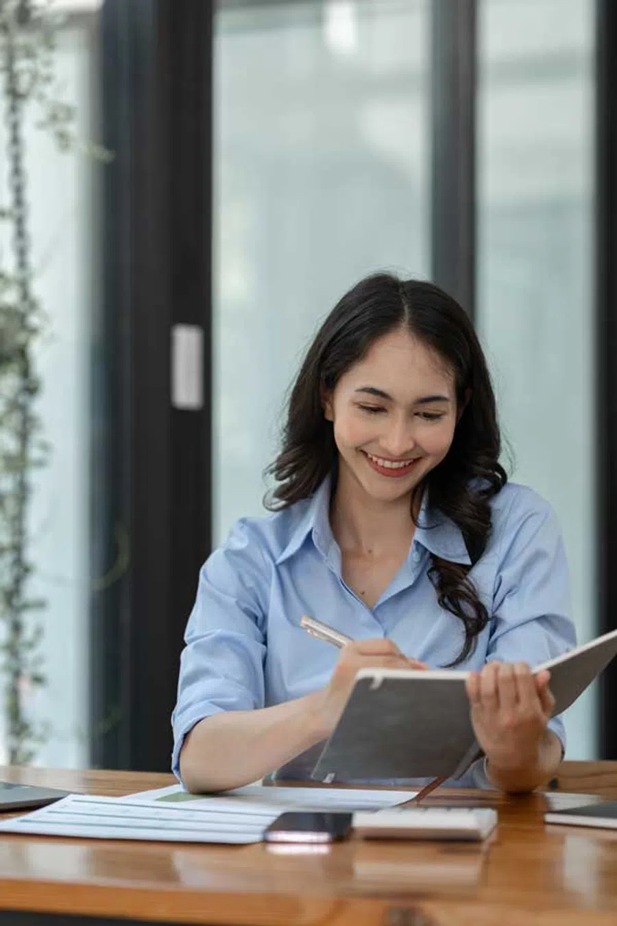 Smiling woman in blue shirt writing in a notebook at a wooden desk with papers and phone.