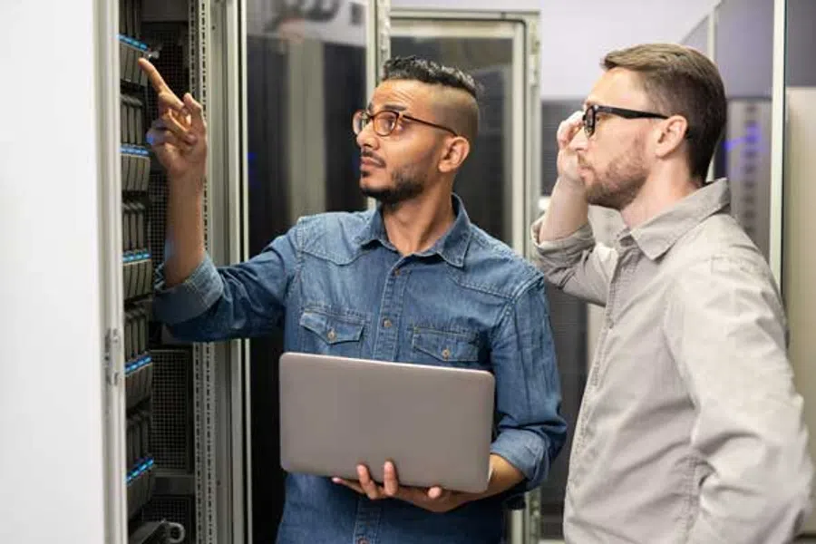 Two IT professionals analyzing server racks while one holds a laptop and points at hardware components