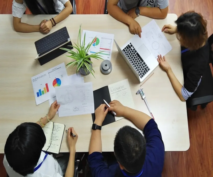 Top view of a team meeting with diverse people discussing charts, notes, and laptop around a table.