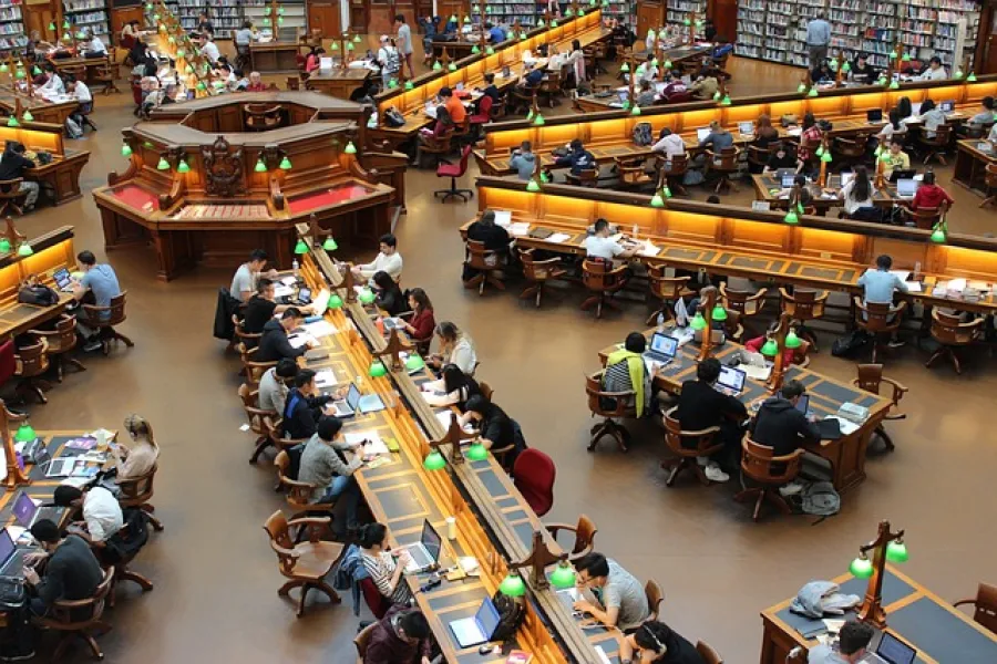 Large library reading room with many people studying at wooden desks and green desk lamps under warm lighting.