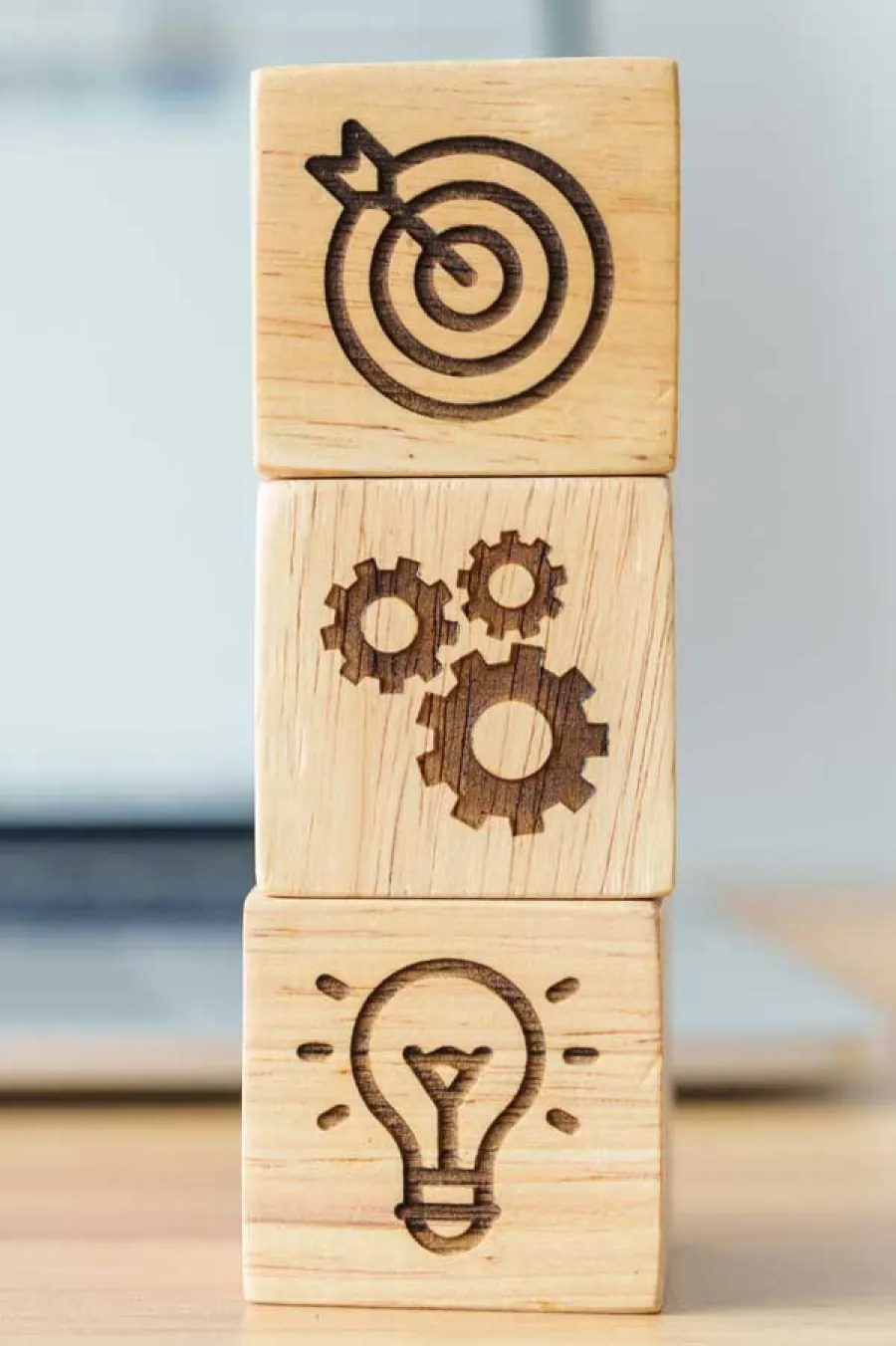Stacked wooden blocks with engraved icons of a lightbulb, gears, and target with arrow on a desk background