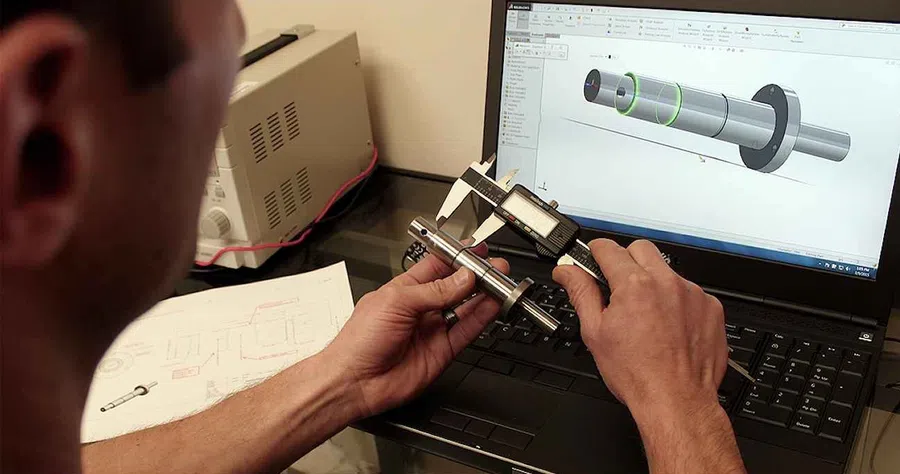 Engineer measures a metal part using digital caliper comparing it to 3D CAD model on laptop screen