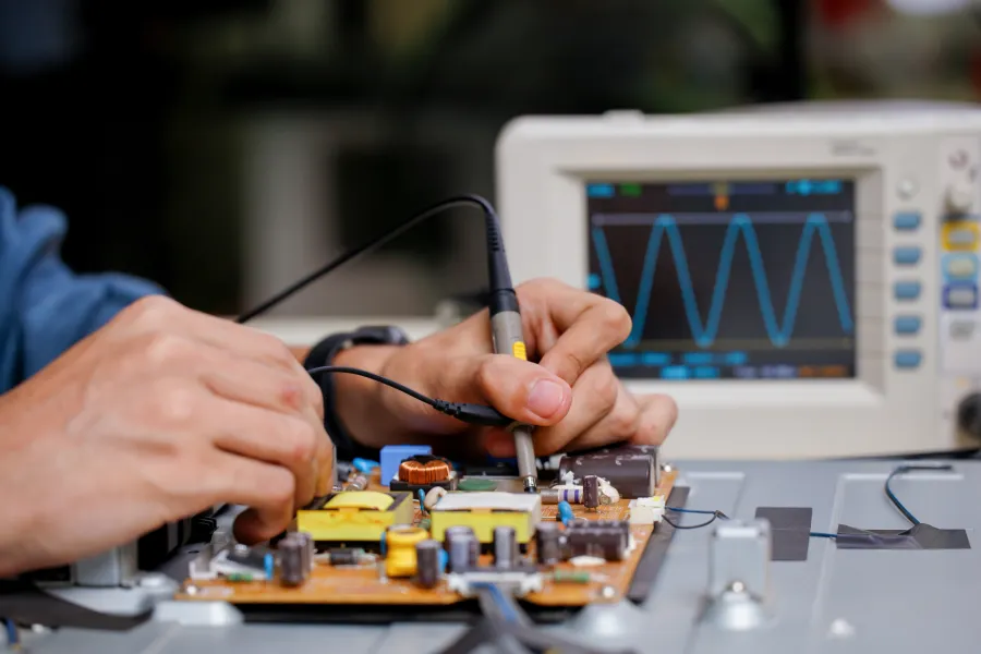Technician testing electronic circuit board with oscilloscope waveform visible on screen in background