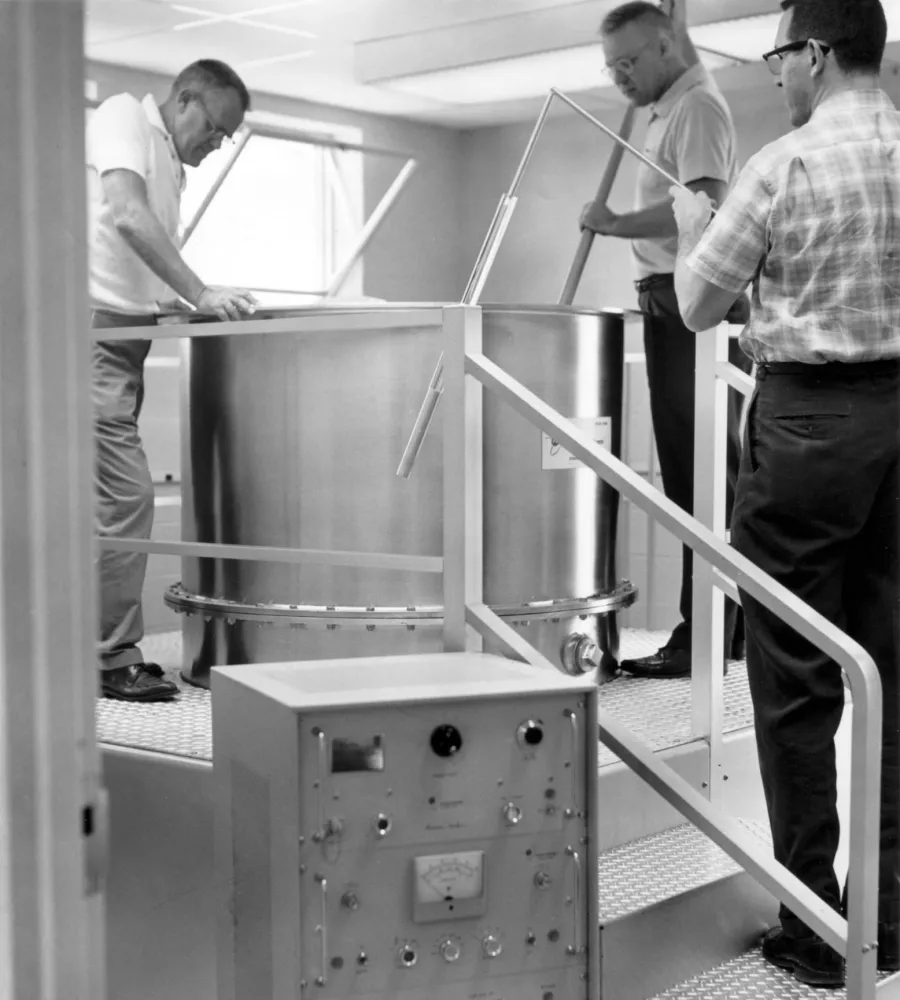 Three men operate and inspect a large metallic industrial machine inside a facility with control equipment nearby.