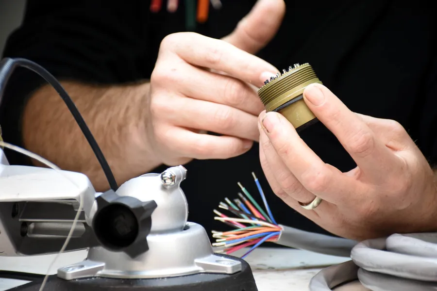 Technician examining a circular metal connector near a multicolored cable bundle on a workbench.