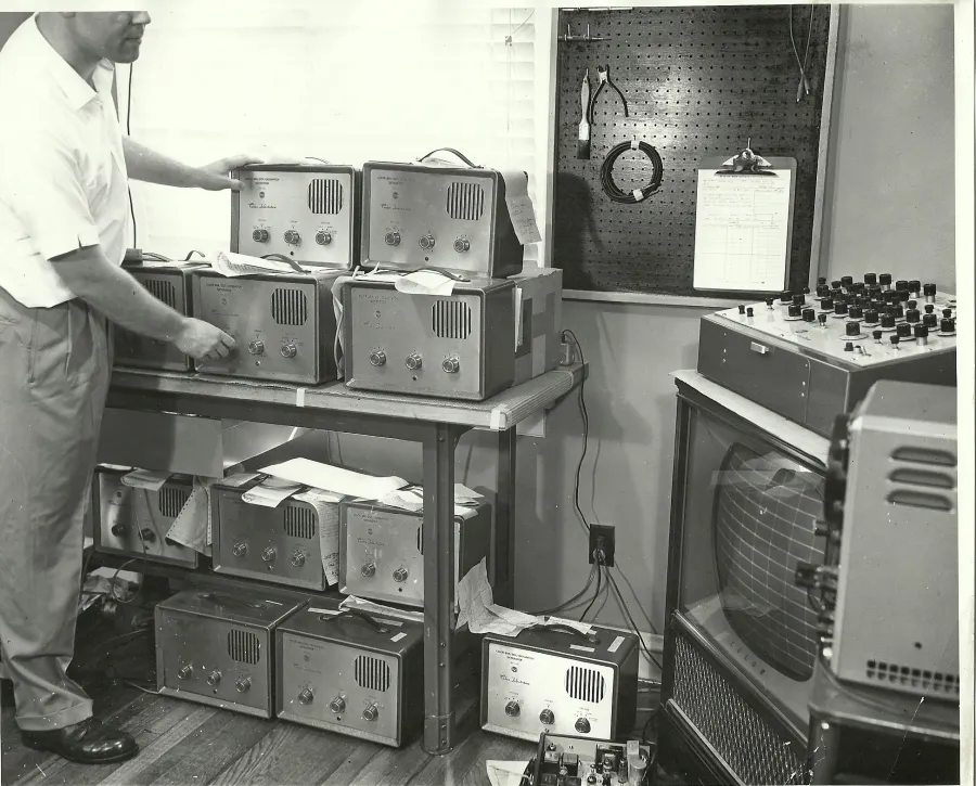 Vintage laboratory with a man adjusting multiple electronic radio or testing equipment on shelves beside an old CRT monitor.
