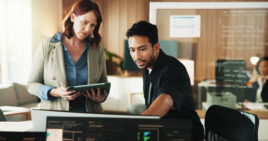 Two professionals collaborating on a tablet and computer screens in a modern office setting with data analysis.