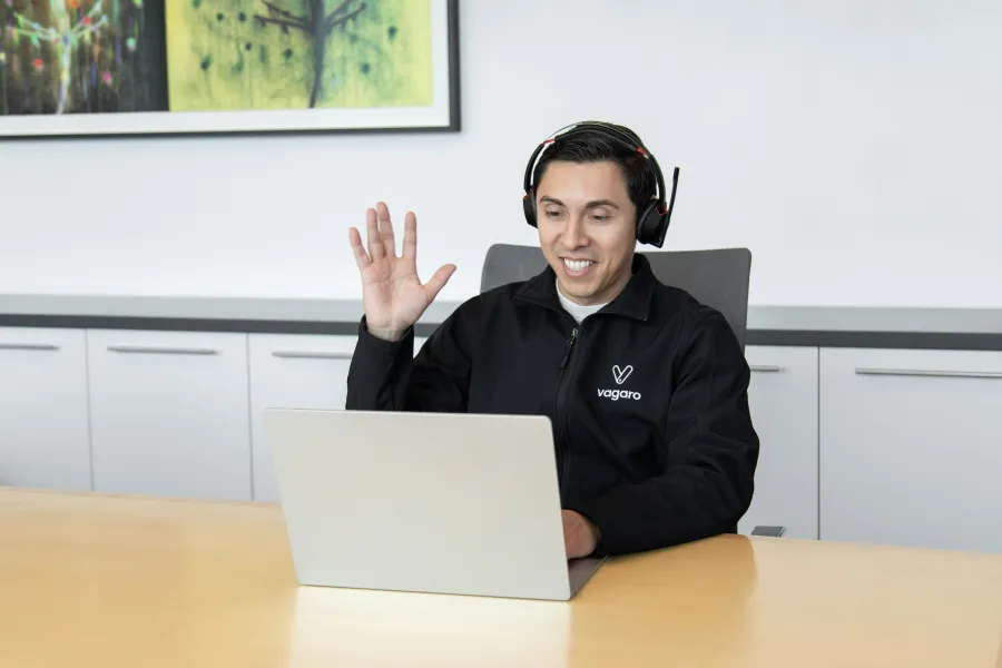Man wearing headphones waves while video chatting on laptop at desk in modern office setting.