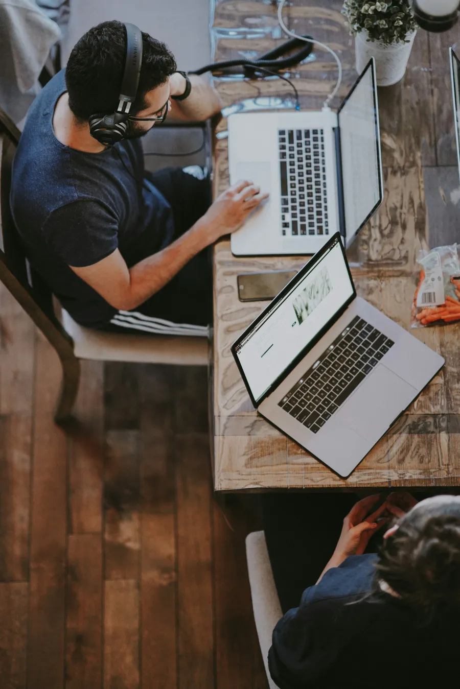 Two individuals working on laptops at a wooden table, headphones on, focused and productive.