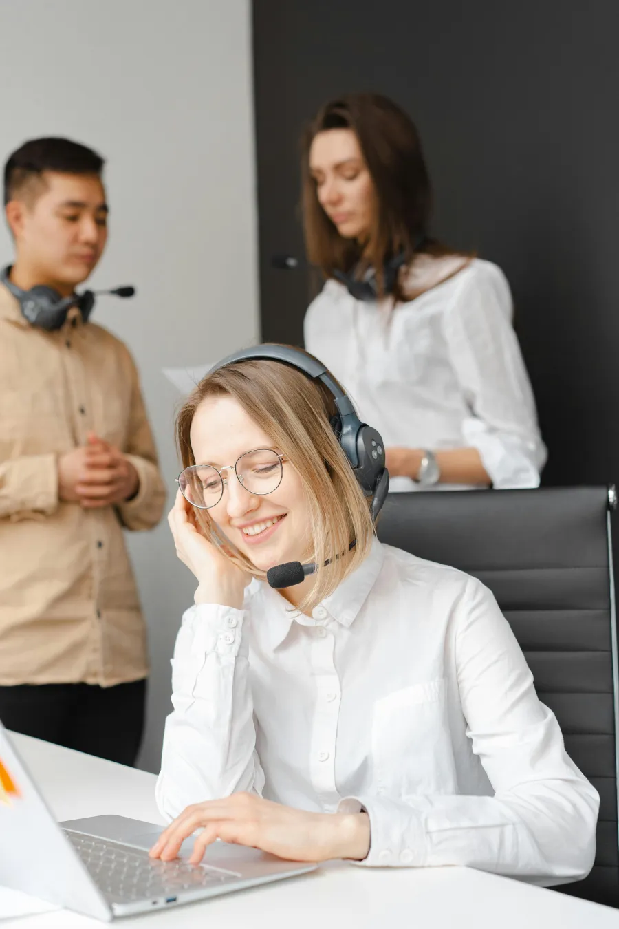 Smiling woman in headset working on laptop with two colleagues in background in modern office setting