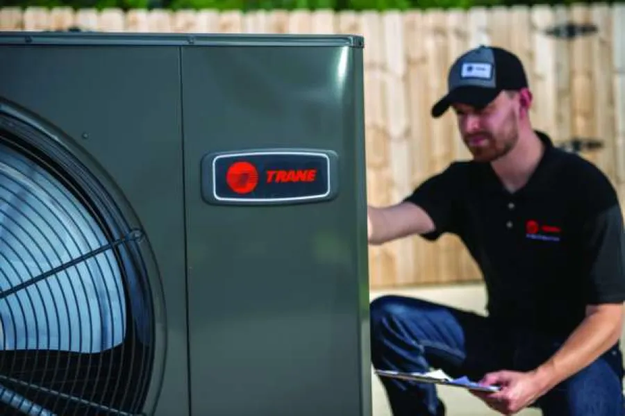 Technician inspecting a Trane HVAC unit outdoors in front of a wooden fence during daytime.
