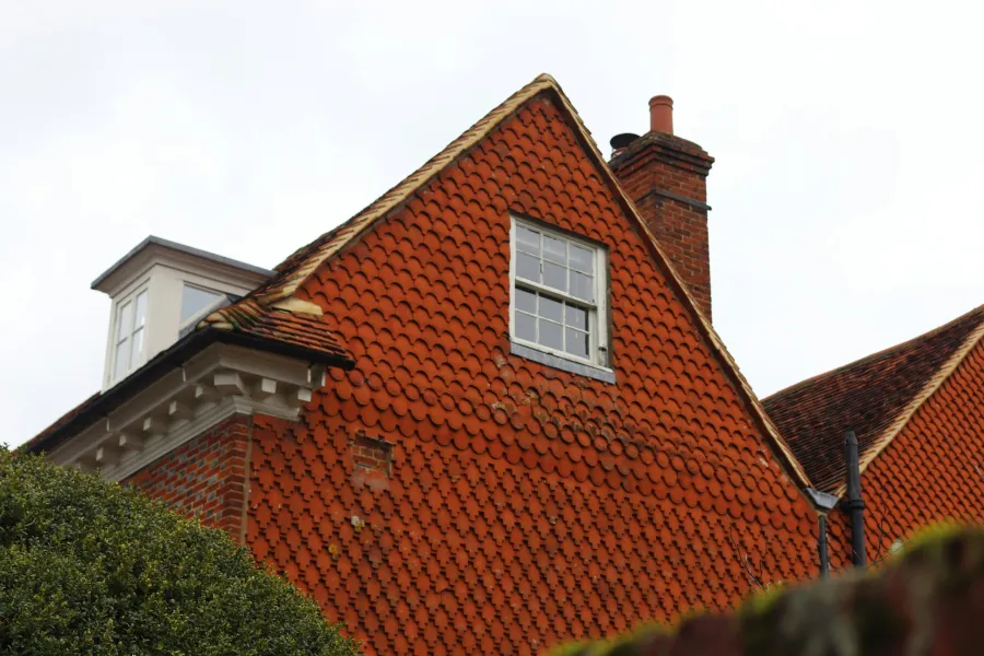 Close-up of a triangular red-tiled roof with a small window and chimney against an overcast sky.