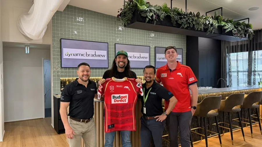 Four men standing in a modern office, one holding a red Budget Direct sports jersey with dynamicbradview screens in the background