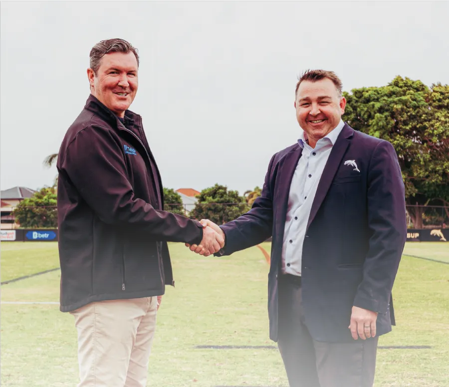 Two men shaking hands outdoors on a sports field, both smiling and dressed in business casual attire.