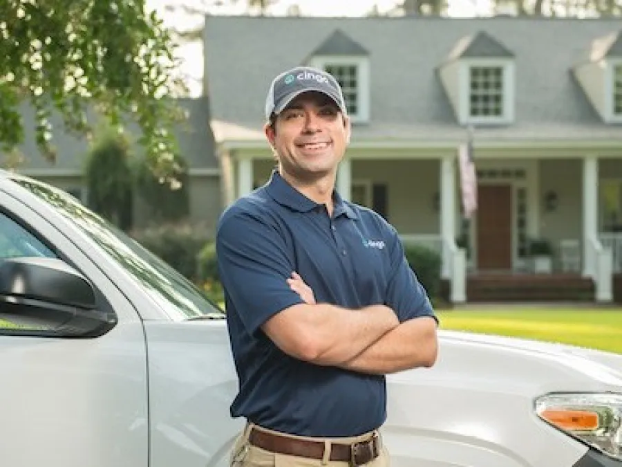 Home service technician in uniform standing with arms crossed by a company vehicle in front of a suburban house.